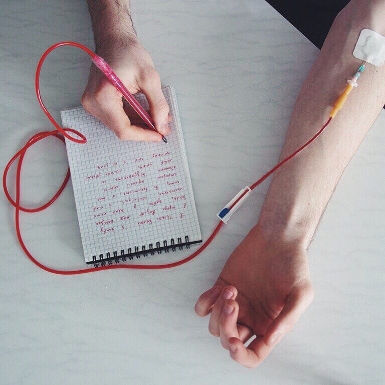 A person's hand holding a pink pen writing in a notebook, with an IV line connected to the arm and a syringe attached.